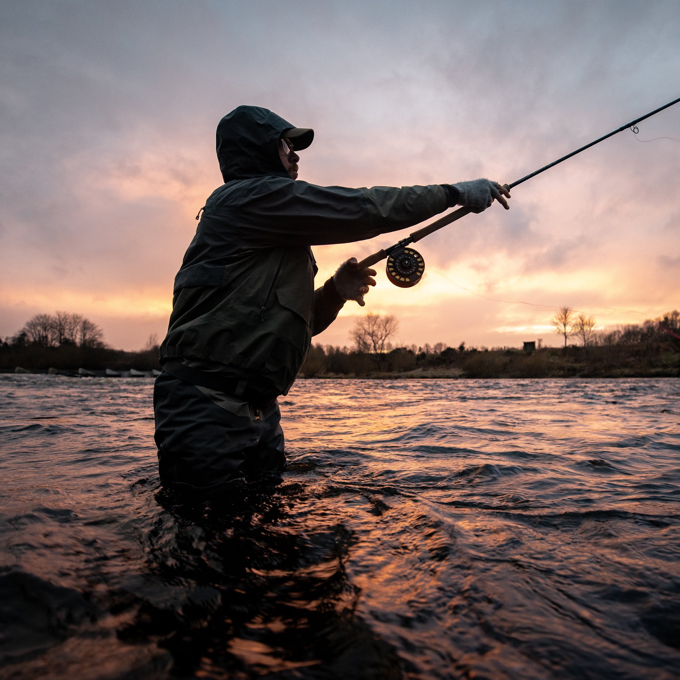 A person in dark waterproof clothing stands in shallow water at sunset, casting a fishing rod across the frame. The scene is silhouetted against a cloudy sky with warm orange and pink light on the horizon and reflections on the water.