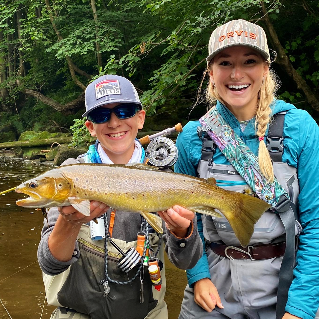 Two people holding a large fish by a river with fishing gear.
